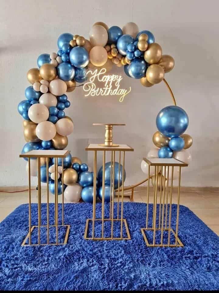 A luxurious birthday decoration setup featuring a golden circular arch adorned with a cascade of metallic blue, gold, and white balloons. A white 'Happy Birthday' neon sign glows in the center, with three elegant golden plinths of varying heights placed in front on a plush royal blue carpet.