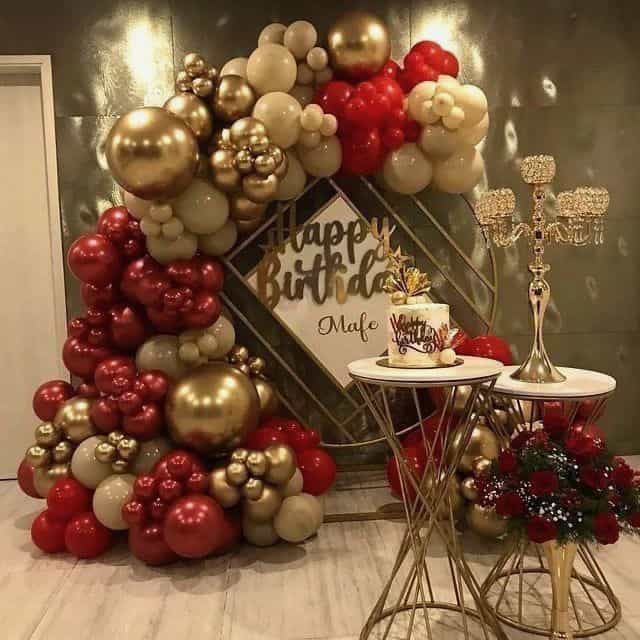 A luxurious birthday decoration with a large gold and red balloon arch, a "Happy Birthday Mafe" sign, an elegant cake, and a candelabra, perfect for a romantic birthday decor.