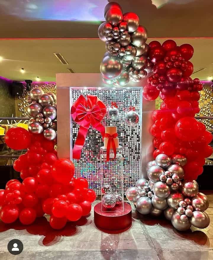 A vibrant red and chrome silver organic balloon garland frames a sparkling silver sequin backdrop with a large red bow, display pedestals, and festive accents.