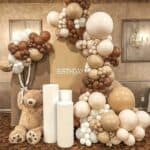 An elegant indoor birthday decoration with an arch-shaped backdrop featuring the word "BIRTHDAY". The setup includes a large organic balloon garland in shades of brown, beige, and white, a oversized teddy bear, and three white cylindrical pedestals, all placed on a patterned carpet against a textured wall with framed artwork and a chandelier.