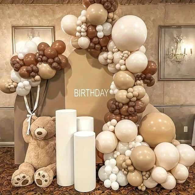 An elegant indoor birthday decoration with an arch-shaped backdrop featuring the word "BIRTHDAY". The setup includes a large organic balloon garland in shades of brown, beige, and white, a oversized teddy bear, and three white cylindrical pedestals, all placed on a patterned carpet against a textured wall with framed artwork and a chandelier.