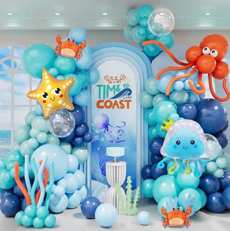 An elaborate under-the-sea themed birthday decoration with balloon arches in shades of blue and aqua. The setup features large foil balloons of an orange octopus, a yellow starfish, a blue jellyfish, and small orange crabs, centered around a backdrop that reads "Time to Coast."