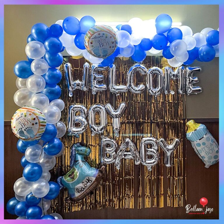 A vibrant Welcome Baby Boy decoration featuring a balloon arch of blue and white balloons, silver foil letters spelling "WELCOME BOY BABY," and a gold fringe backdrop. Two round foil balloons with "Welcome Baby Boy" text, a baby bottle balloon, and a rocking horse balloon are also part of the display.