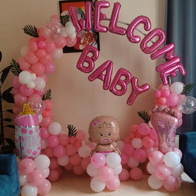 A festive pink and white balloon arch decoration for a baby shower or "Welcome Baby Girl" event. The decoration includes a large pink foil "WELCOME BABY" banner, a balloon arch with pink and white balloons, a baby-shaped foil balloon, a bottle-shaped foil balloon, and a large foot-shaped "It's a Girl!" foil balloon.