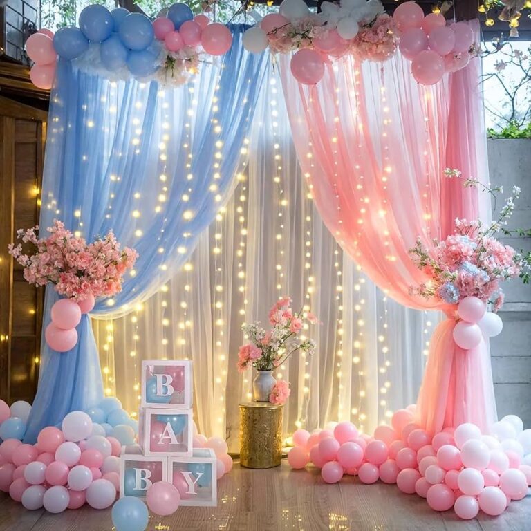 A stunning baby shower or gender reveal backdrop features sheer blue and pink draped curtains adorned with string lights, creating a soft, glowing effect. Balloon garlands in matching pink, blue, and white cascade over the top and frame the sides. Floral arrangements in pink and blue are placed on the curtains and on either side of the display. Clear, block-like boxes spelling out "B-A-B-Y" and clusters of pink, blue, and white balloons fill the foreground and the floor.