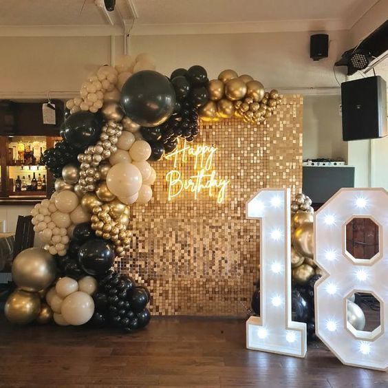 A lavish 18th birthday party decoration setup featuring a gold sequin shimmer wall backdrop with a neon "Happy Birthday" sign. An enormous organic balloon garland in black, gold, and cream drapes over the wall. To the right, large, light-up marquee numbers "18" stand on a dark wooden floor.